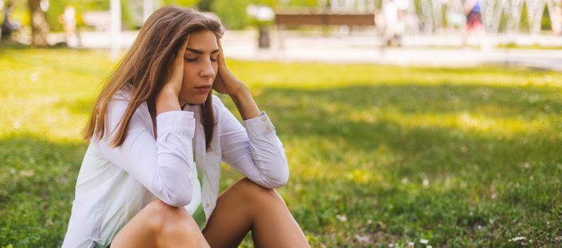 A photo of a woman sitting in the grass with a severe headache holding her head in her hands.