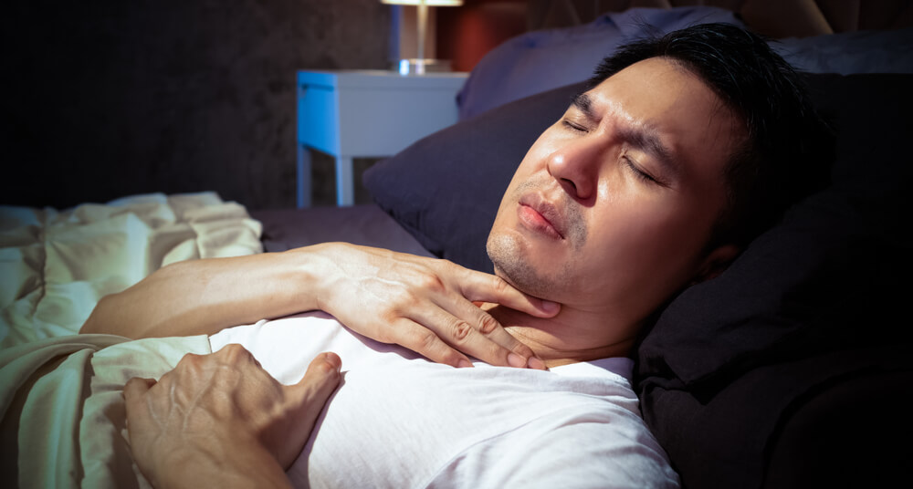 A man is lying down in bed grabbing his throat with pain from acid reflux.