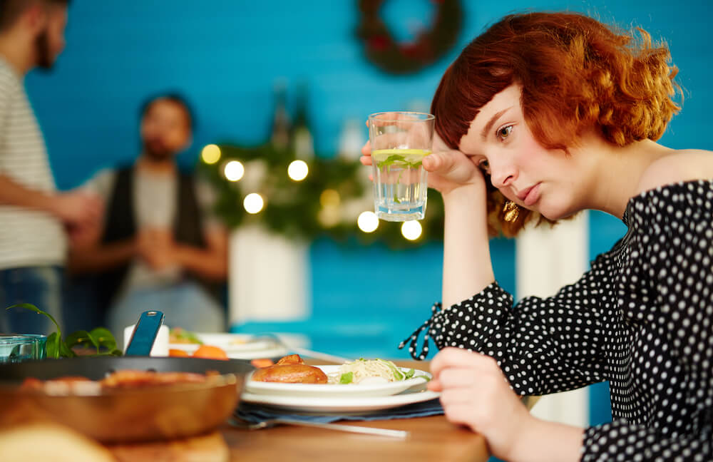 A woman sits at a table eating lunch while holding her glass of water near her head.