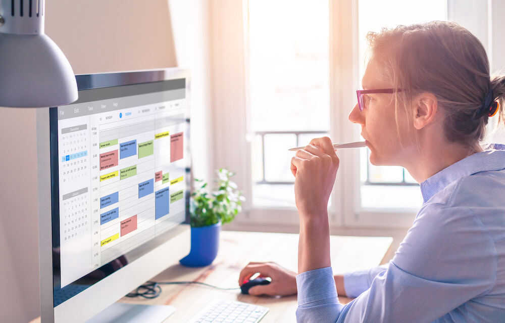 A woman is sitting at her desk while looking at her calendar on her computer.