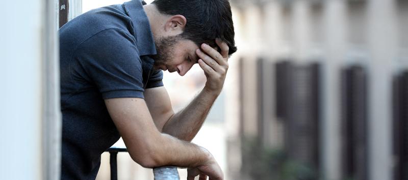 A photo of a man leaning on the railing of a balcony while holding his forehead. 