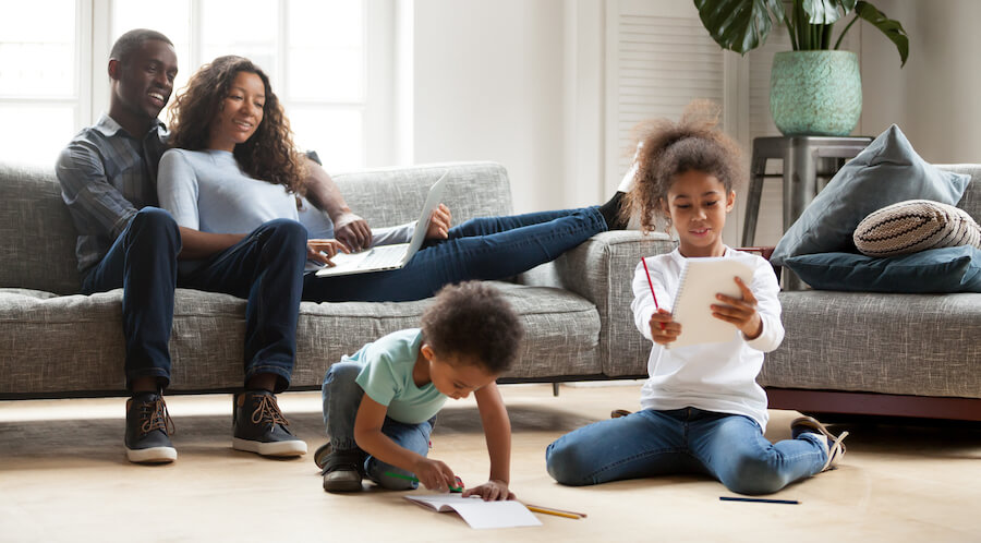 A mom and dad are sitting on the couch working and looking at their children drawing and playing on the floor. 