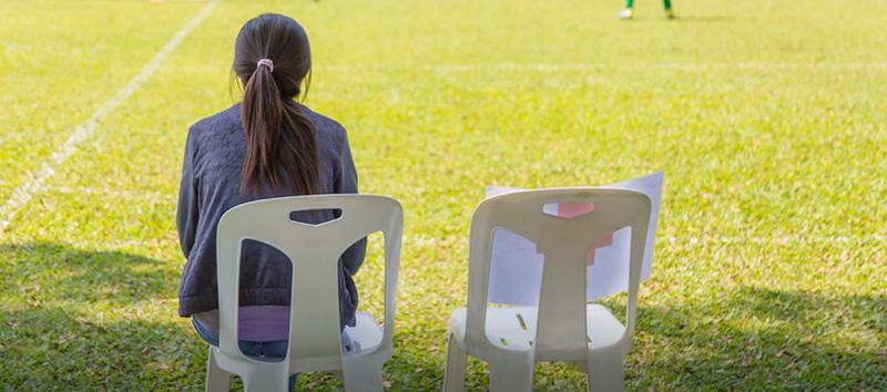 A woman is sitting in a chair watching a football game with an empty seat next to her.