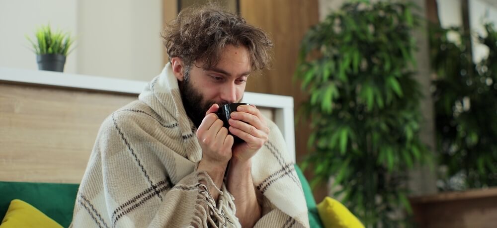 A man is sick sitting on the couch drinking hot tea.