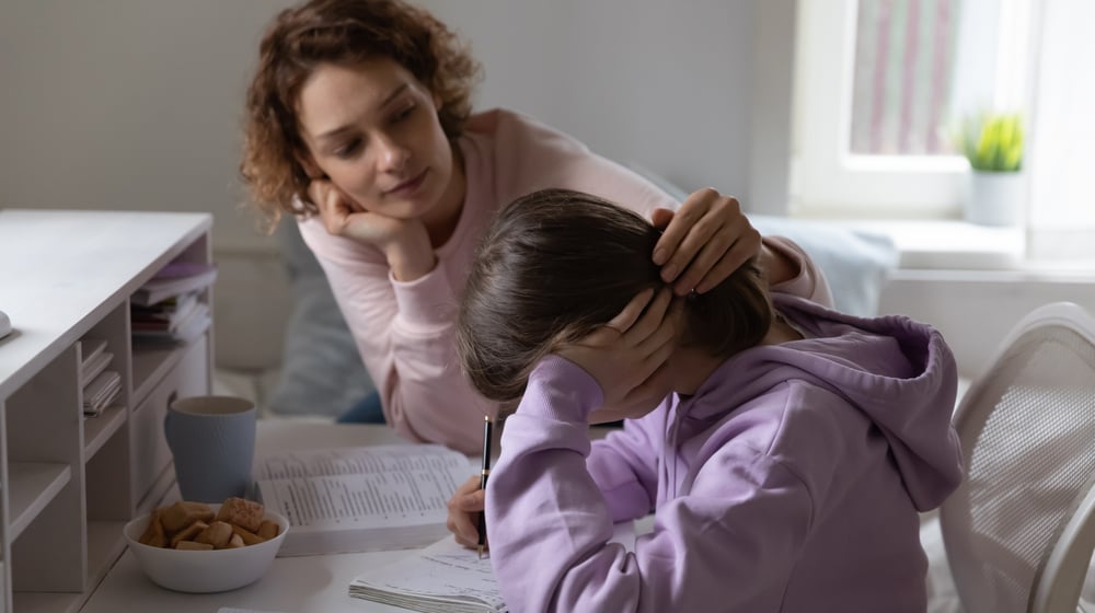 A mom puts her hand on the back of her daughters head while she's doing her homework.