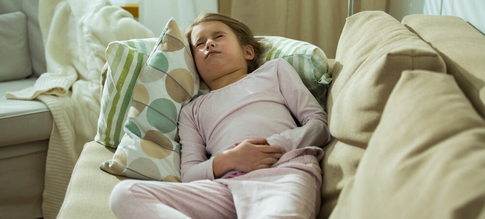 A young girl is lying on the couch while grabbing her stomach in pain.