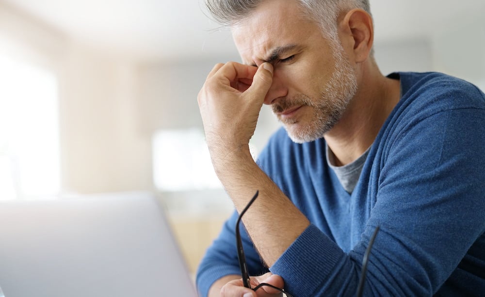 Man sitting down at his desk while holding the bridge of his nose with one hand and his reading glasses with the other.