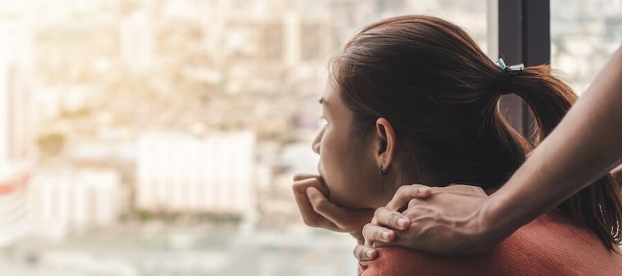 A woman stares pensively out the window with her hand on her chin and someone else's hand on her shoulder.