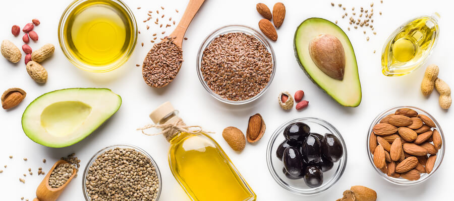 A photo showing olive oil, avocados, olives, and different types of nuts laid out on a table.