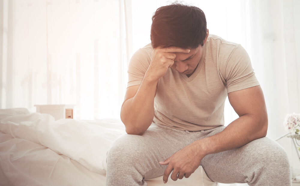 A man is sitting on his bed with his head in his hand.