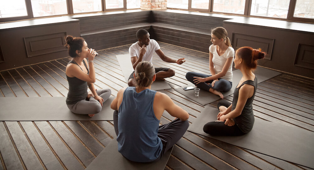 A group of 5 people sitting on the floor before their yoga class.