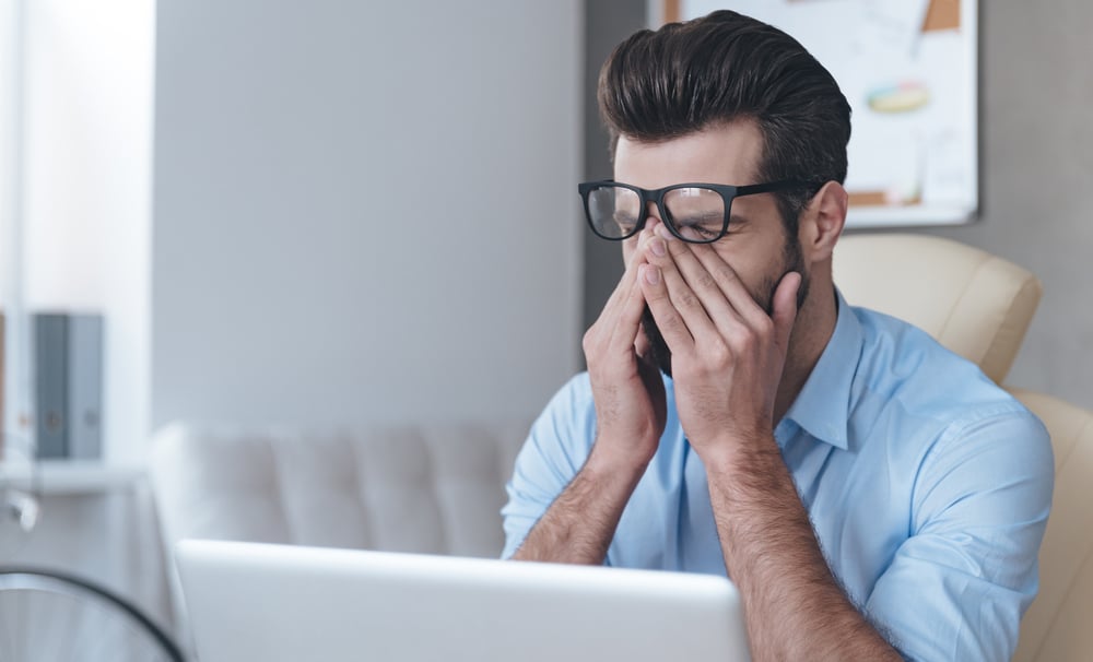 A man pushes his glasses up while rubbing his eyes in front of his computer.
