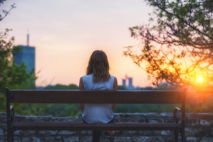 A young woman sitting on a bench during golden hour while staring at the cityscape.