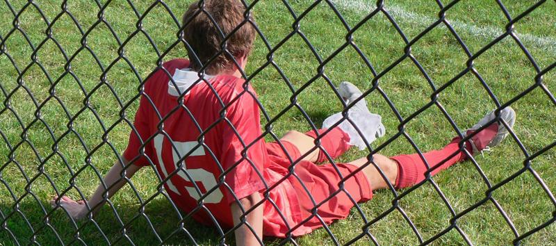 A young boy is sitting on the field behind a fence wearing his jersey.