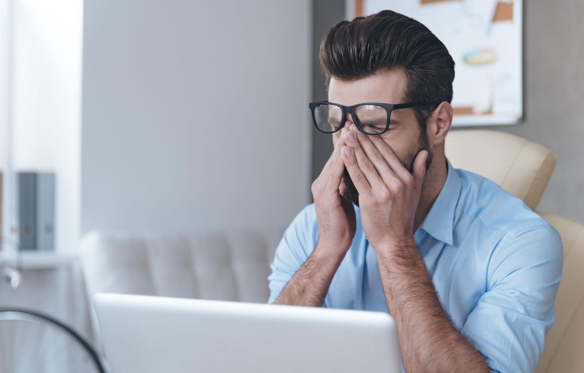 A tired man is rubbing his eyes while working in his home office