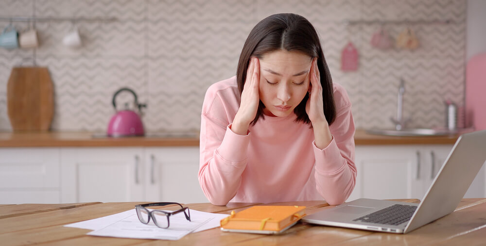 A woman is grabbing her head while sitting at her desk in the kitchen